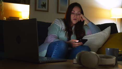 Mujer-Sentada-En-Un-Sofá-En-Casa-Por-La-Noche-Transmitiendo-O-Viendo-Películas-O-Programas-En-Una-Computadora-Portátil-Y-Navegando-Por-Internet-En-El-Teléfono-4